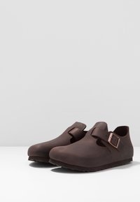Brown rubber clogs with an open design, featuring a single strap and metallic buckle. Smooth texture and rounded toe shape. Non-slip sole.
