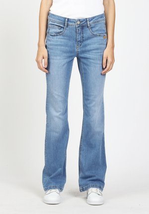 Hellblaue Bootcut-Jeans, getragen mit weißen Sneakers und einem weißen Oberteil, gezeigt von der Taille bis zu den Füßen vor einem neutrale Hintergrund.