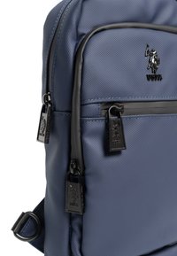 Námořnický modrý batoh se strukturovanou tkaninou, dvěma zipovými kapsami a černými detaily. Vlastnosti zahrnují logo a kontrastní švy.