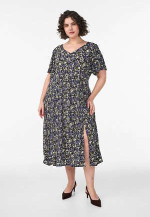 Žena v čiernych kvetovaných midi šatách s bočným rázporkom a krátkymi rukávmi, v spojení s čiernymi lodičkami na podpätku so zapínaním na pätu, stojaca na bielom pozadí.