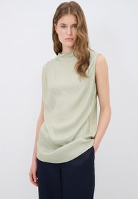 Top senza maniche di colore verde chiaro, realizzato in tessuto morbido, presenta un collo alto e un design drappeggiato, abbinato a pantaloni blu navy scuri.