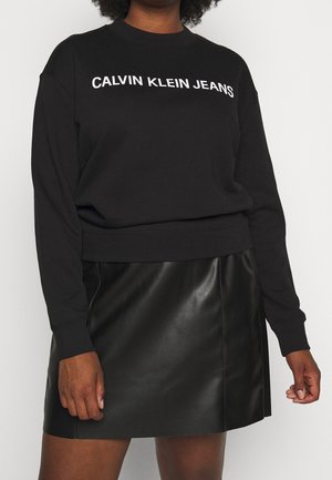 Osoba ubrana w czarną bluzę Calvin Klein Jeans z długim rękawem i czarną skórzaną spódnicę, stojąca na tle jednolitego tła.