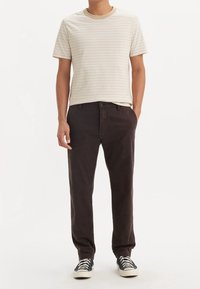 T-shirt à rayures beige avec un col rond, associé à un pantalon marron à jambe droite. Des baskets noires avec des semelles blanches complètent le look.