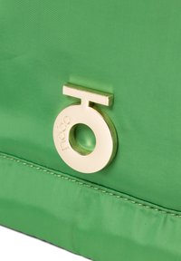 Vihreä nailonilaukku, jossa on sileä pinta, pyöreä kultainen logo-aksentti ja suorakulmainen solki. Minimalistinen suunnittelu, puhtaat linjat.