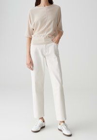 Beige gestricktes Oberteil mit rundem Halsausschnitt und aufgeschlagenen Ärmeln, kombiniert mit weißen geraden Hosen und weißen Sneakers mit braunen Akzenten.