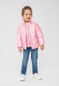 Veste rose gonflée avec une texture matelassée brillante, fermeture éclair et col montant, portée sur une chemise rose clair et un jean bleu. Baskets blanches.