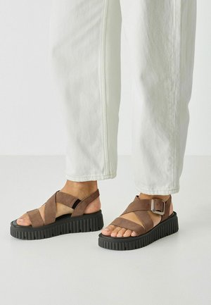 Brown suede sandals with crisscross straps, a buckle detail, and a thick black platform sole, paired with light-colored cropped pants.