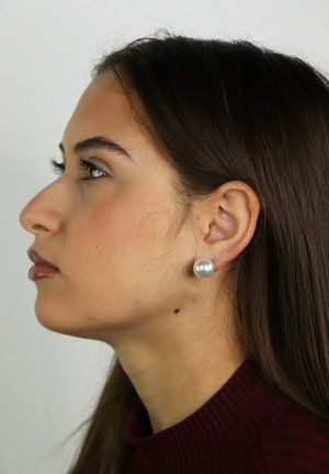 Femme aux longs cheveux foncés et à la boucle d'oreille en perle, portant un haut bordeaux, vue de profil gauche sur un fond clair uni.