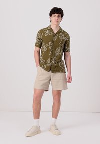 Short-sleeve, olive green shirt with white floral patterns, paired with light beige shorts. Model wears white socks and cream sneakers.