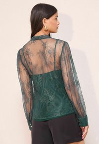 Gennemsigtig, mørkegrøn blondebluse med lange ærmer, blomster mønstre, en figursyet silhuet og knappede manchetter. Blød tekstur med elegant detaljering.