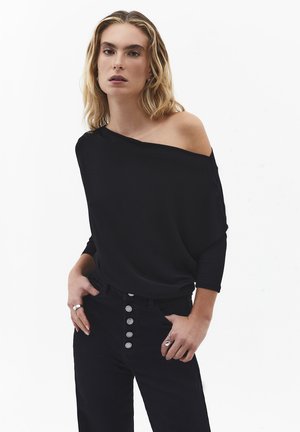 Blondynka w czarnej bluzce z odkrytymi ramionami i rękawami do łokci oraz czarnych, wysokich spodniach z pięcioma guzikiami, stojąca z rękami w kieszeniach.