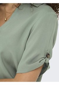 Blusa a maniche corte, verde chiaro, con scollo a V, caratterizzata da un dettaglio con polsini abbottonati e una texture liscia e morbida.