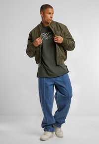 Olivengrøn bomberjakke med ribbede manchetter, lagd over en mørkegrøn T-shirt, parret med løse blå denimjeans og lyse sneakers.