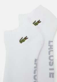 Chaussettes blanches en coton jusqu'à la cheville avec un logo de crocodile brodé vert et le texte "LACOSTE" en gris sur les côtés. Bord ribbé pour un maintien optimal.
