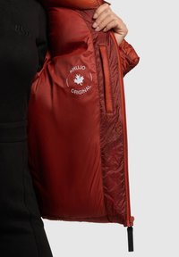 Wnętrze czerwonej pikowanej kurtki o gładkiej fakturze; zawiera nadrukowane logo "KHUJO ORIGINAL" oraz kieszeń na zamek po boku.