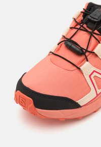 Trail running shoe with a coral textile upper, black rubber sole, and a secure lacing system. Features reinforced toe cap and padded collar.