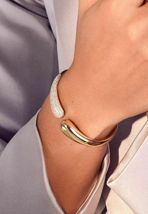 Guldmetall cuff armbånd med et glat, buet design, akkompagneret af et afsnit af klare krystaller for ekstra detaljer og tekstur.