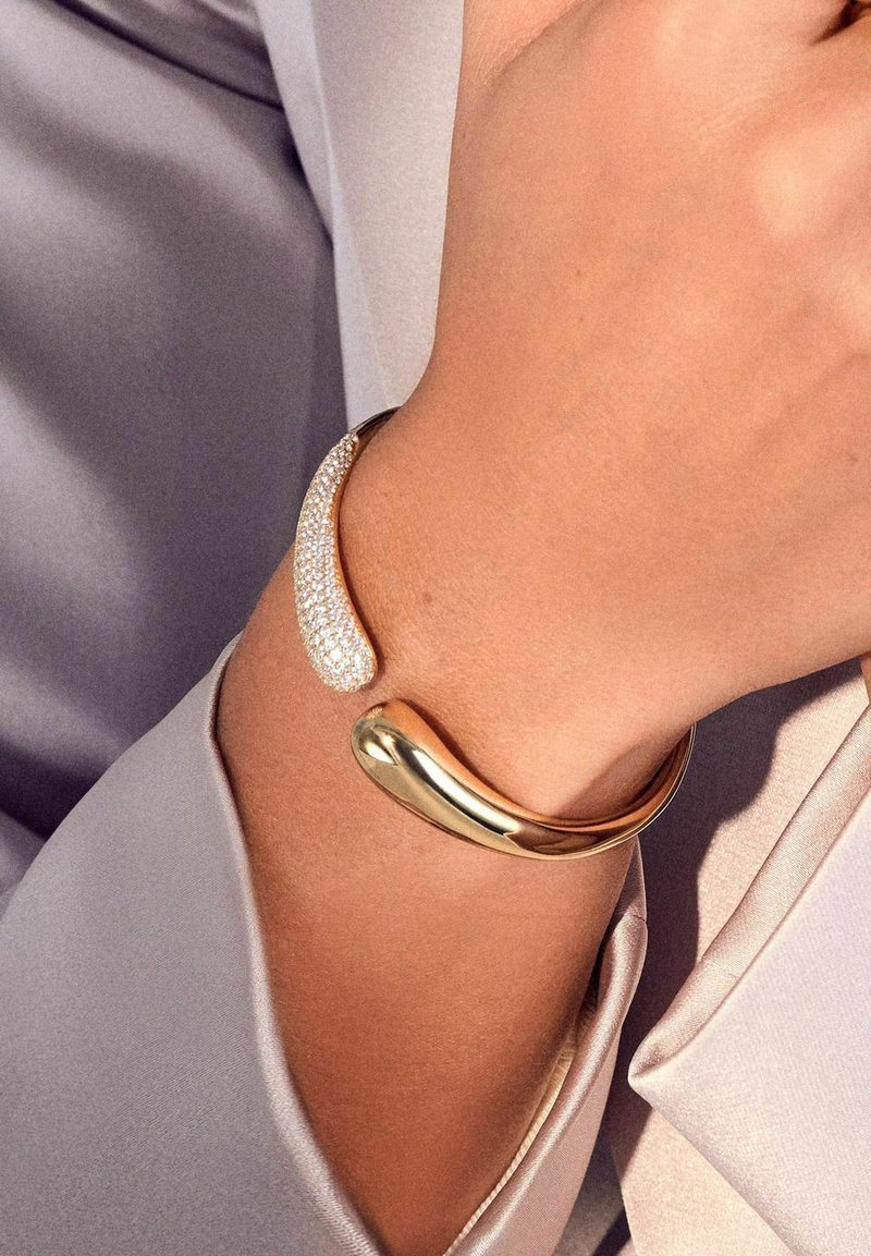 Bracciale in metallo dorato con un design liscio e curvo, arricchito da una sezione di cristalli trasparenti per un tocco di dettaglio e texture.