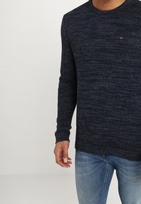 Marinblå stickad tröja med ett texturerat mönster, rund halsringning och långa ärmar. Har en liten logotyp på bröstet.