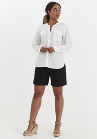 Hvid, letvægts langærmet skjorte med krave, to brystlommer og knaplukning foran. Sammen med sorte shorts og beige wedges.