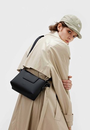 Sort, struktureret taske med klaplukning og justerbar rem. Fremstillet af glat materiale, båret over kroppen. Beige trenchcoat i baggrunden.