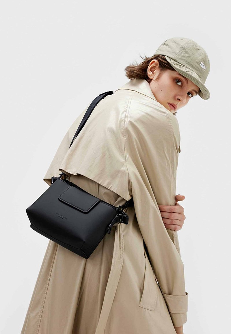 Sort, struktureret taske med klaplukning og justerbar rem. Fremstillet af glat materiale, båret over kroppen. Beige trenchcoat i baggrunden.
