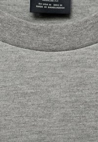 Tejido de punto gris de una camiseta de cuello redondo. Presenta un ajuste regular con una textura suave. La etiqueta indica la talla y el origen.