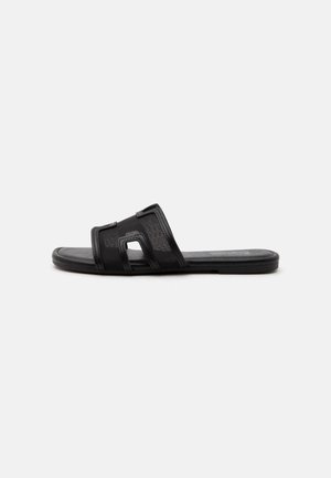 Sort flad slidesandal med en bred netrem med udskårne geometriske former på en ensfarvet hvid baggrund.