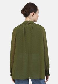 Olive green blouse with a textured dot pattern, button closure at the back, and long sleeves, made from a lightweight, sheer fabric.