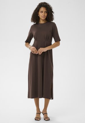 Femme aux cheveux bouclés portant une robe midi marron à manches courtes, fente latérale, et des sandales noires à talons, debout sur fond blanc.