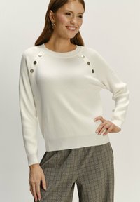 Biała sweter z żeberkowym wzorem, z guzikami na ramionach i długimi rękawami, w połączeniu z spodniami w kratę. Miękka tekstura i dopasowany krój.