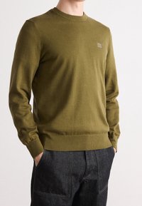 Pull tricoté vert olive avec un col rond, des poignets et un ourlet côtelés ; présente une petite étiquette de marque à l'avant. Porté sur un pantalon foncé.