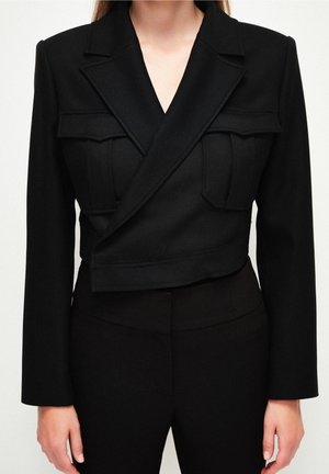 Nainen, jolla on päällään istuva musta bleiseri, jossa on jämäkät olkapäät, kaksi suurta etutaskua ja mustat, korkeavyötäröiset housut, jotka sopivat bleiseriin.