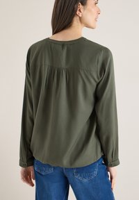 Blouse à manches longues vert olive avec un col rond, dotée d'un empiècement plissé au dos, portée avec un jean bleu et des manches retroussées.