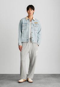 Világoskék farmerdzseki bézs színű gallérral és logóval, fehér póló alatta, szürke melegítőnadrág és krémszínű sportcipő.