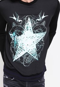 Black sweatshirt featuring a large turquoise star design with a dotted pattern, surrounded by floral elements and birds. Ribbed neckline and cuffs.