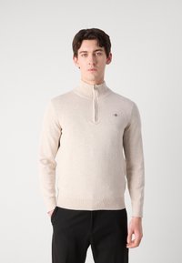 Suéter beige con cremallera en suave tejido de punto, con un alto cuello y puños acanalados. Detalle del logo pequeño en el lado izquierdo del pecho.