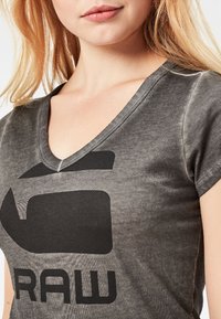 Camiseta gris de cuello en V con un gran logo negro que muestra la palabra "RAW" de manera prominente. Tela suave y ligeramente texturizada, con mangas cortas.