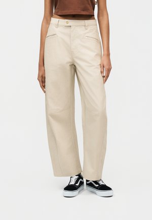 Persoană purtând pantaloni largi bej, adidași alb-negru și un top scurt maro, stând pe un fundal simplu.