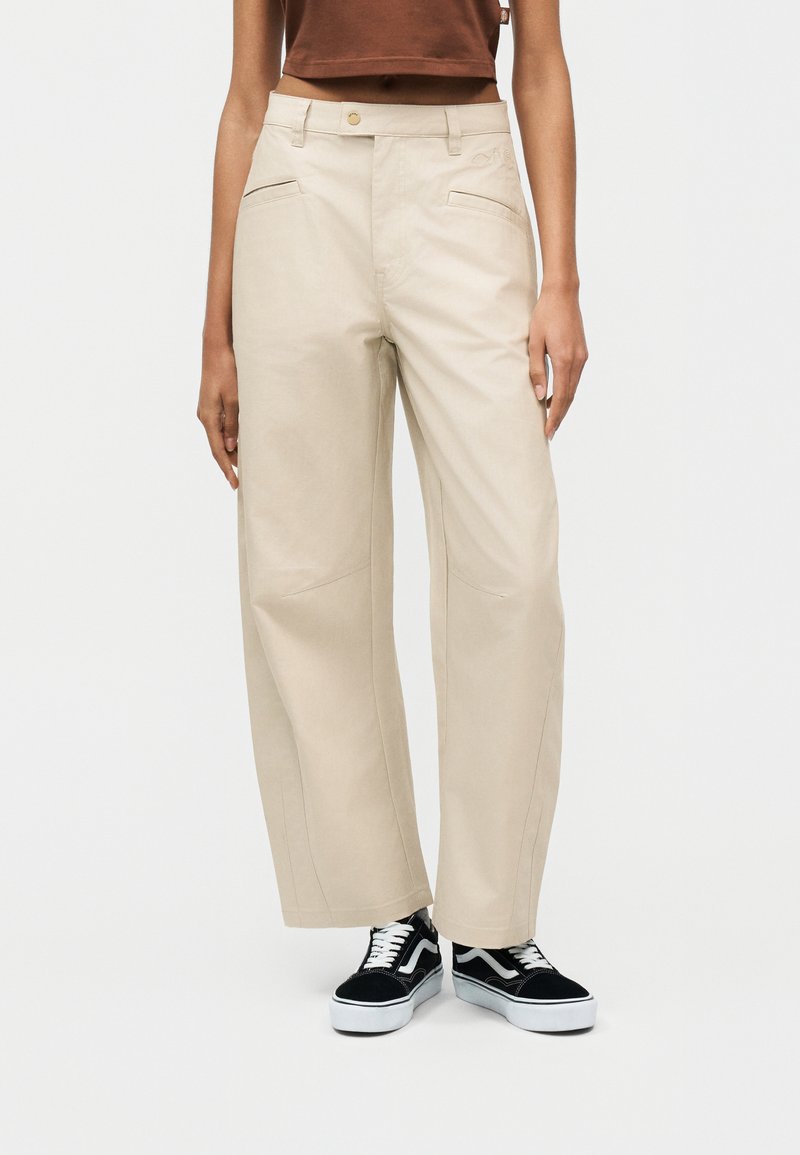 Άτομο που φοράει μπεζ παντελόνι με φαρδύ πόδι, μαύρα και άσπρα αθλητικά παπούτσια και cropped καφέ τοπ, στέκεται μπροστά σε απλό φόντο.