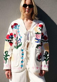 Valge puuvillane jakk, millel on värviline lilleline tikand, ees sidumisvõimalus, kaks taskut, kujundatud erinevate mustrite ja elavate detailidega.