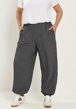 Personne portant un pantalon large à motif quadrillé noir et blanc, des baskets blanches et une chemise blanche unie à manches courtes, les mains dans les poches.