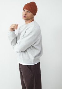 Szary sweatshirt z prążkowanym dołem, noszony na białym T-shircie. W zestawie z ciemnymi brązowymi spodniami i prążkowaną pomarańczową czapką. Model pozuje z boku.