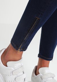 Ciemnoniebieskie obcisłe jeansy z denimu ze złotymi akcentami suwaków przy krawędzi nogawek, zestawione z białymi teksturowanymi sneakersami z paskiem na rzep.