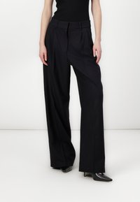 Black pinstriped wide-leg trousers with a high waist, featuring front pleats and smooth fabric texture. Paired with pointed black heels.