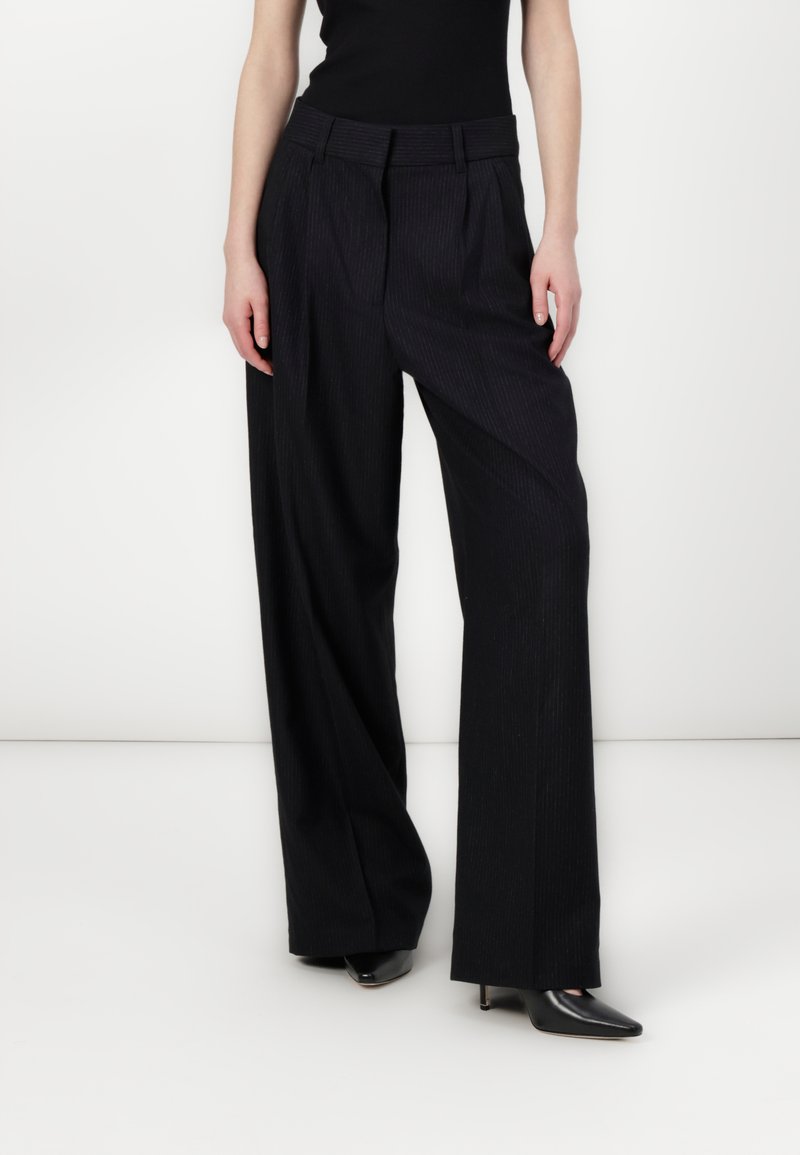 Black pinstriped wide-leg trousers with a high waist, featuring front pleats and smooth fabric texture. Paired with pointed black heels.