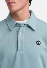 Ljusblå piketröja i mjuk bomull. Har en nedvikt krage, tre knappar och en liten marinblå logotyp på bröstet.