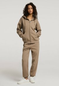 Beżowa bluza z kapturem na zamek, z kieszeniami i sznurkiem, zestawiona z pasującymi, zwężającymi się dresami. Materiał wydaje się miękki; całość dopełniają białe sneakersy.