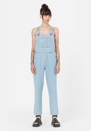 Salopette en denim bleu clair, avec des bretelles ajustables, une poche frontale et des jambes droites, associée à des sandales noires.