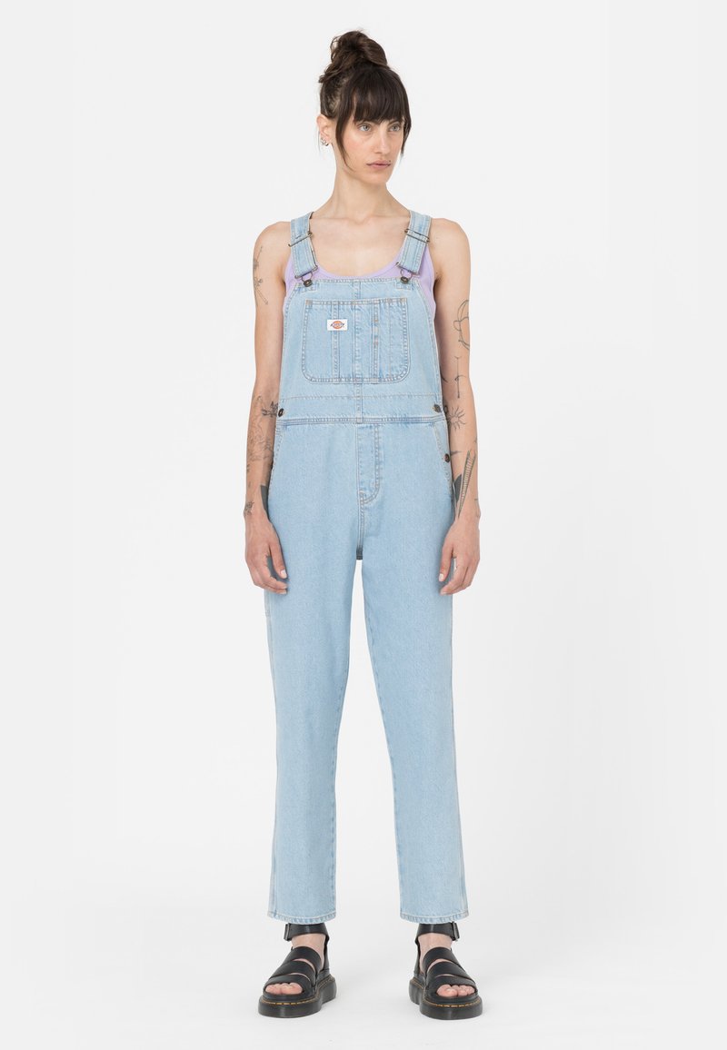 Salopette in denim azzurro chiaro, con spalline regolabili, tasca frontale e gambe dritte, abbinate a sandali neri.